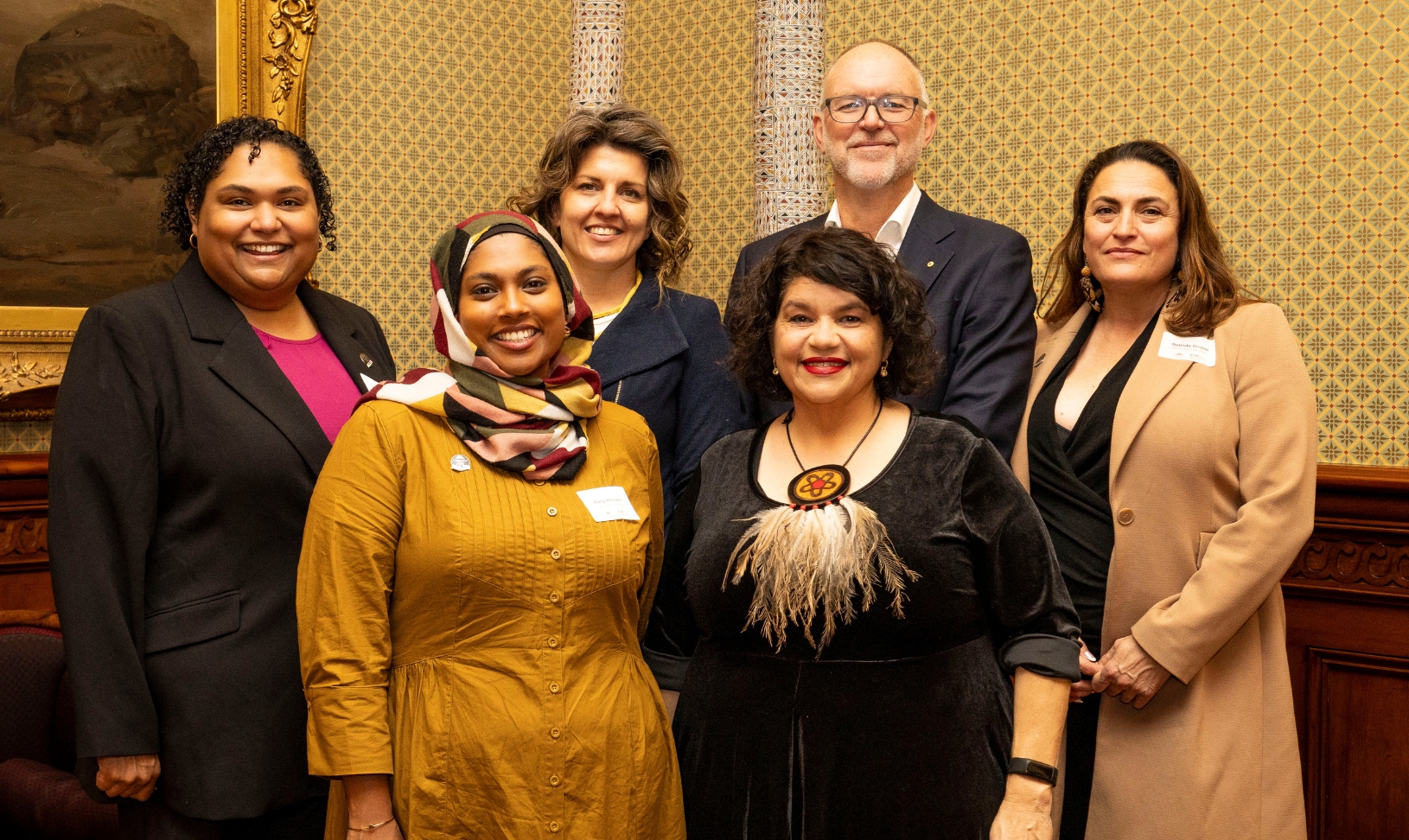 The Reconciliation Australia Board - from left to right, Stacee Ketchell (dark skin, short curly hair, black suit over pink shirt), Rana Hussain (dark skin, hijab with a black, white, yellow and ochre headscarf and a long yellow dress), Liz Brennan (light skin, wavy dark hair with light highlights, blue suit), Kirstie Parker (light skin, shoulder-length black hair, large necklace with woven design, black dress), Michael Rose (white, tall, bald with white beard, glasses and charcoal suit, white shirt with no tie) and Belinda Duarte (light skin, dark brown hair, tan long coat over black top). They are standing together in a stately room, with an artwork in an elaborate gold frame visible behind them.