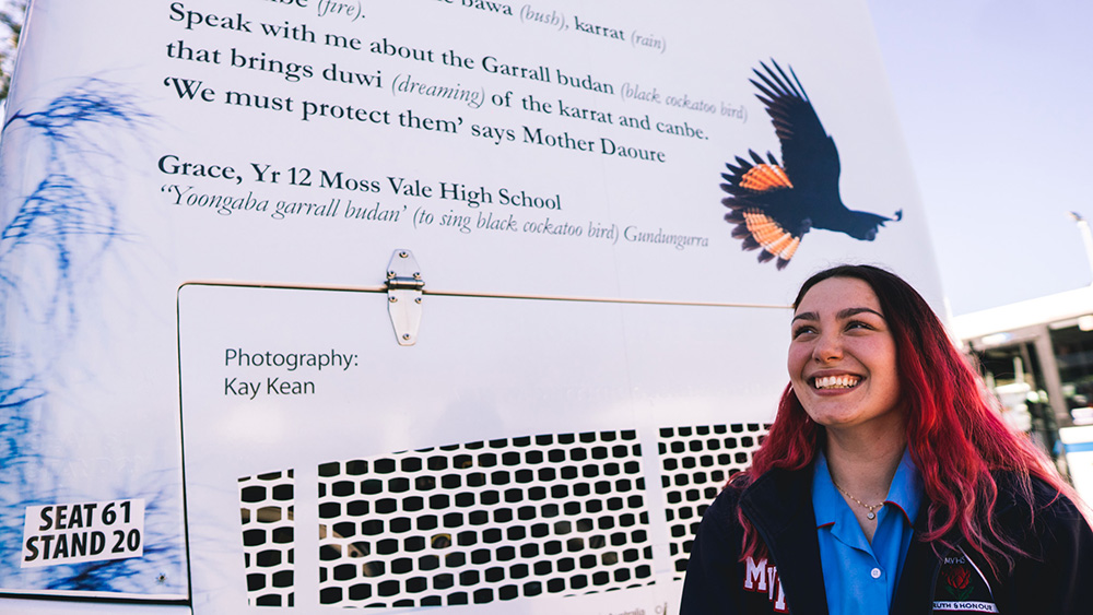 A woman smiling as she stands behind a bus that displays First Nations language