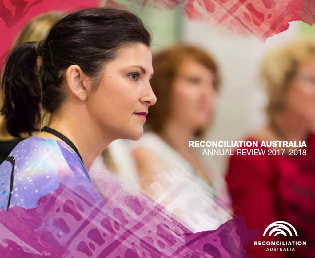 Cover of Reconciliation Australia Annual Review 2017-2018. Image shows the side profile of a woman in the foreground, with other people out of focus behind her. She looks ahead, as if listening to someone speak to the group