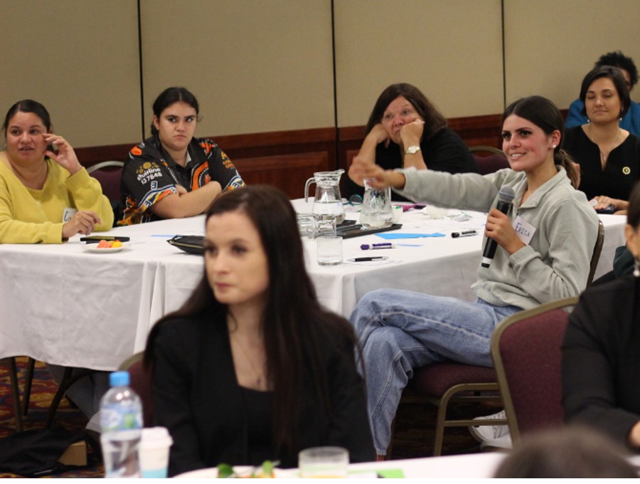 Young women at a conference table and one is talking into a microphone