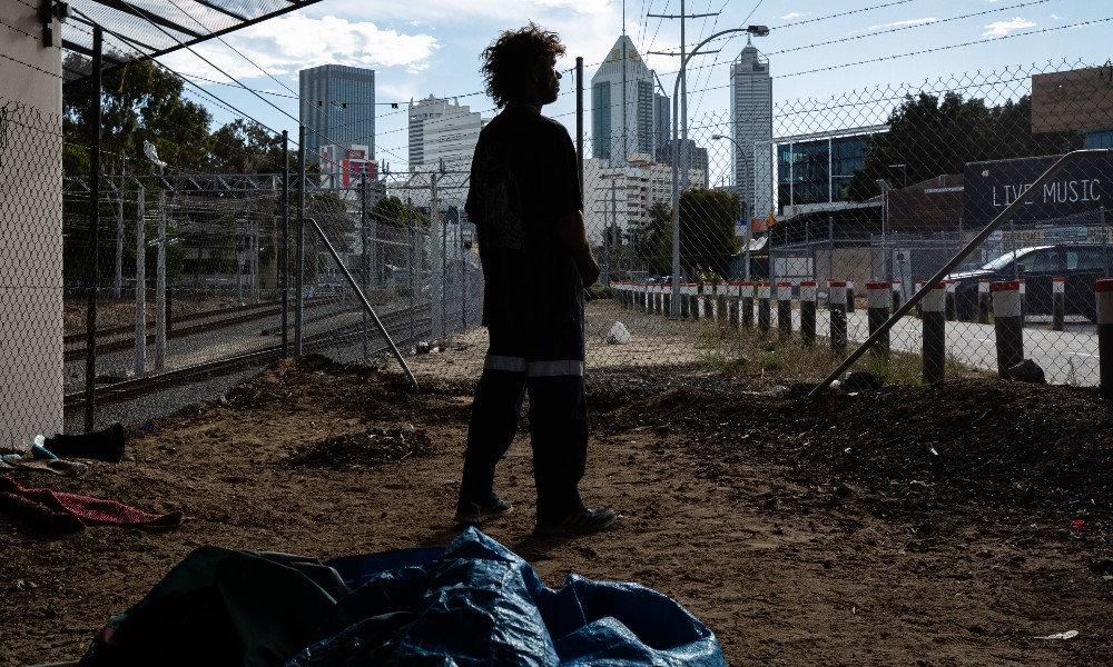 A person experiencing homelessness stands above their sleeping bag under a bridge in Perth.
