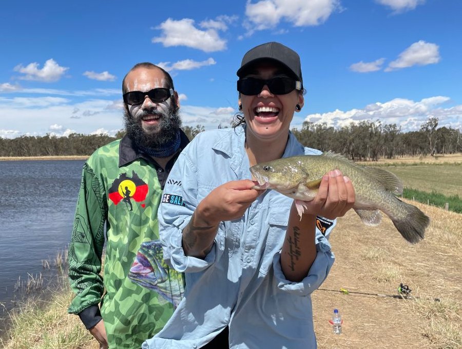 A young man and a young woman. Both are smiling and laughing. The woman is holding a fish she has just caught.