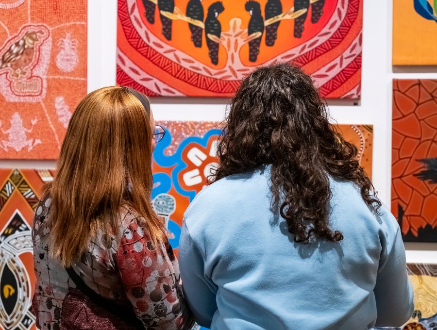 Two women look at various artworks on a wall.