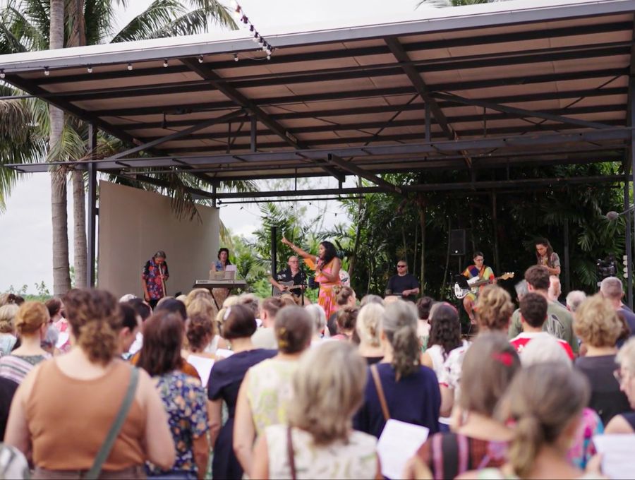 A large crowd stands in front of a stage in the sun, being conducted by a woman standing on the stage.