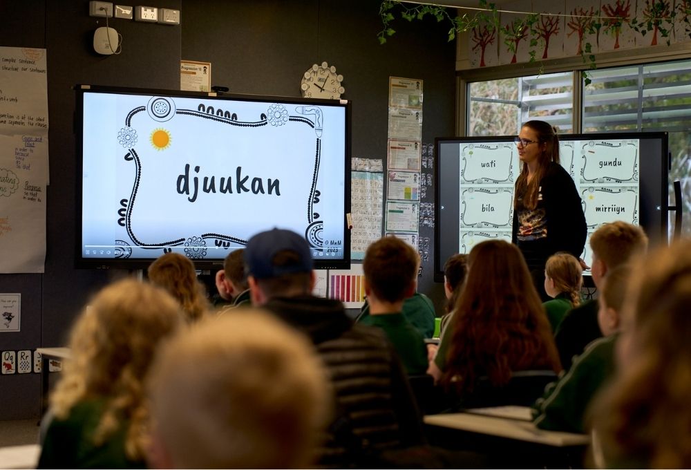 A group of students sit in front of a screen with the word 'djuukan' on it.
