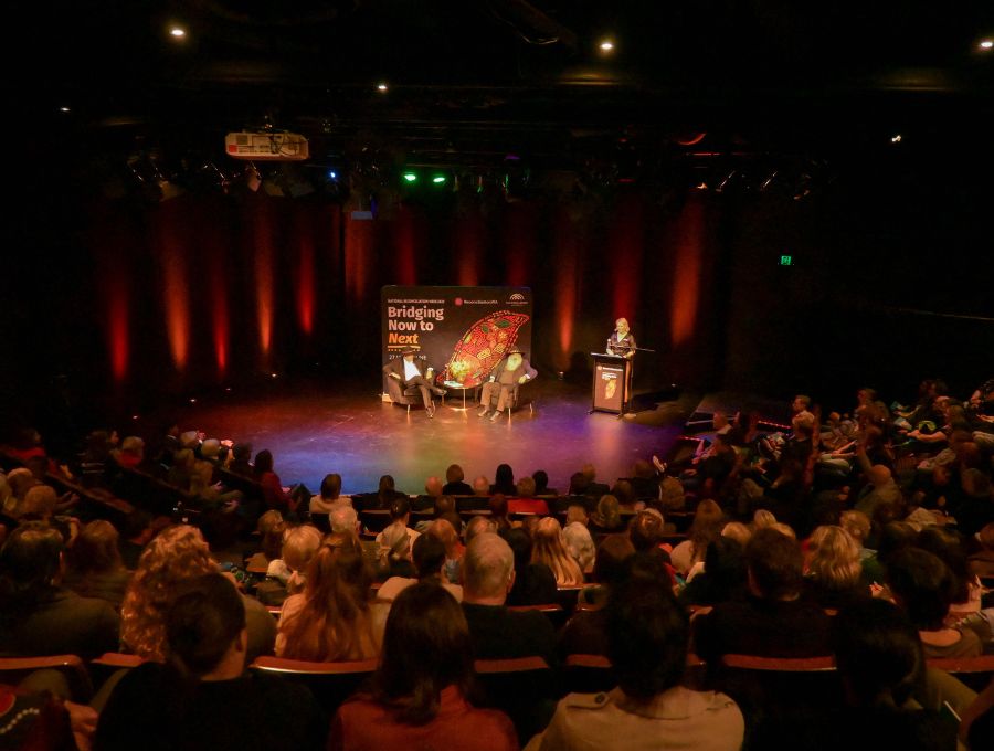 A large crowd watches two men talking on stage.
