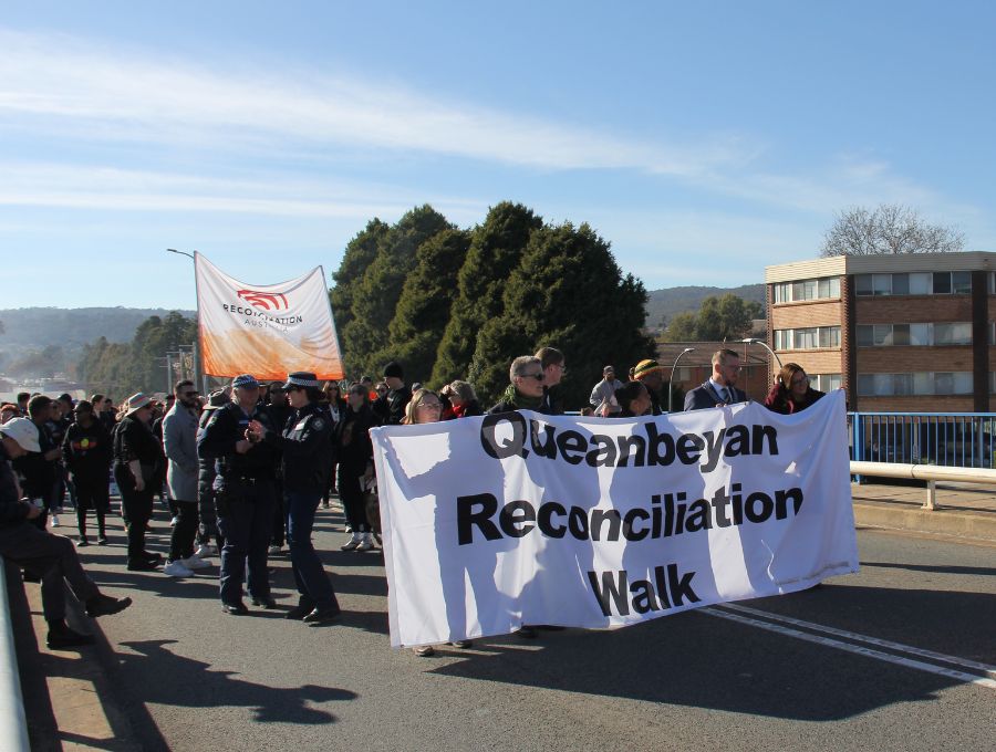 A crowd walks along a road. There is a banner at the front reading 'Queanbeyan Reconciliation Walk', and another behind that says 'Reconciliation Australia'.