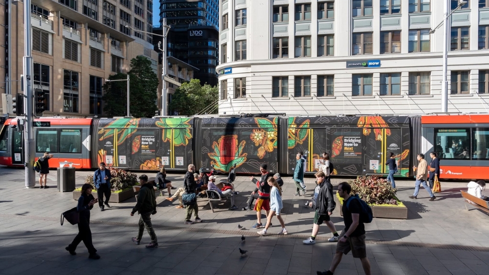 A group of people walking past a tram in Sydney with the NRW 2025 artwork on it.