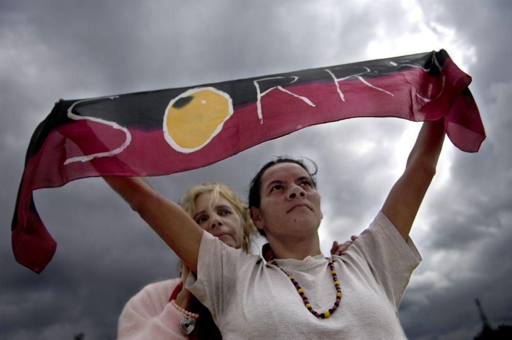 Woman with dark hair holding a banner painted red, black and yellow that reads 'SORRY' with a blonde woman standing behind her.