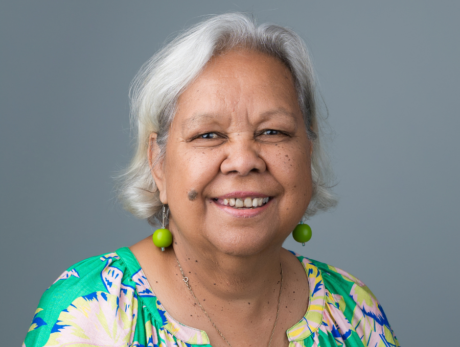 An elderly Aboriginal woman, smiling, wearing green earrings and a green floral-patterned dress.