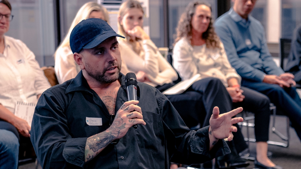 A man in a cap and a button-shirt with tattoos on his arms speaks into a microphone in front of a group of people.