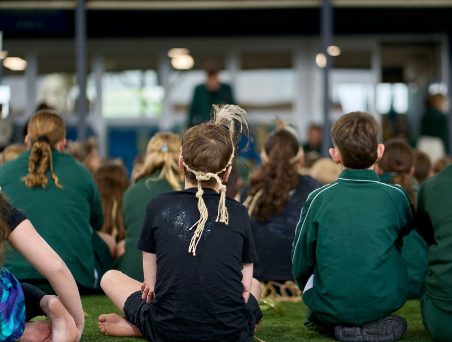 A group of schoolchildren seen from the back, watching an assembly. The child in the middle is wearing a traditional headdress and a t-shirt with ochre stains visible.