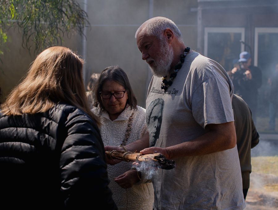 An Elder in a t-shirt that reads 'Mannalargenna Day' shows a woman a smoking stick.