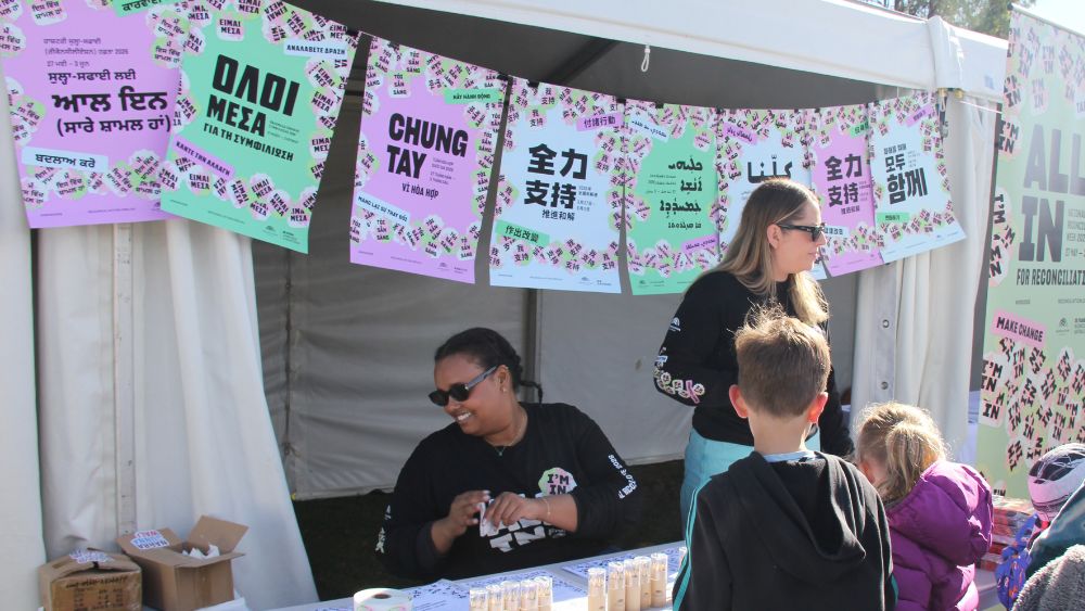 Two women in NRW 2026 ALL IN t-shirts, standing in front of a string of multilingual NRW 2026 posters with their distinctive blue, pink and green pastel colourway, speak to a group of children at an outdoor event stall.