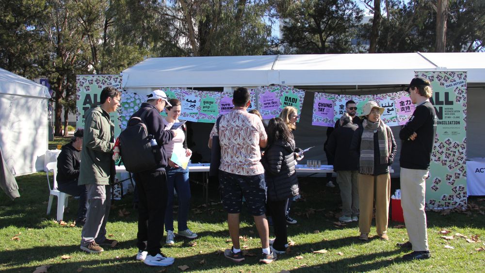 A group of people standing in front of an event stall on a sunny day. The stall is decorated with pink, green and blue pastel colours of the NRW 2026 campaign, and the theme, ALL IN, is visible on branding, as are multilingual versions of the poster.