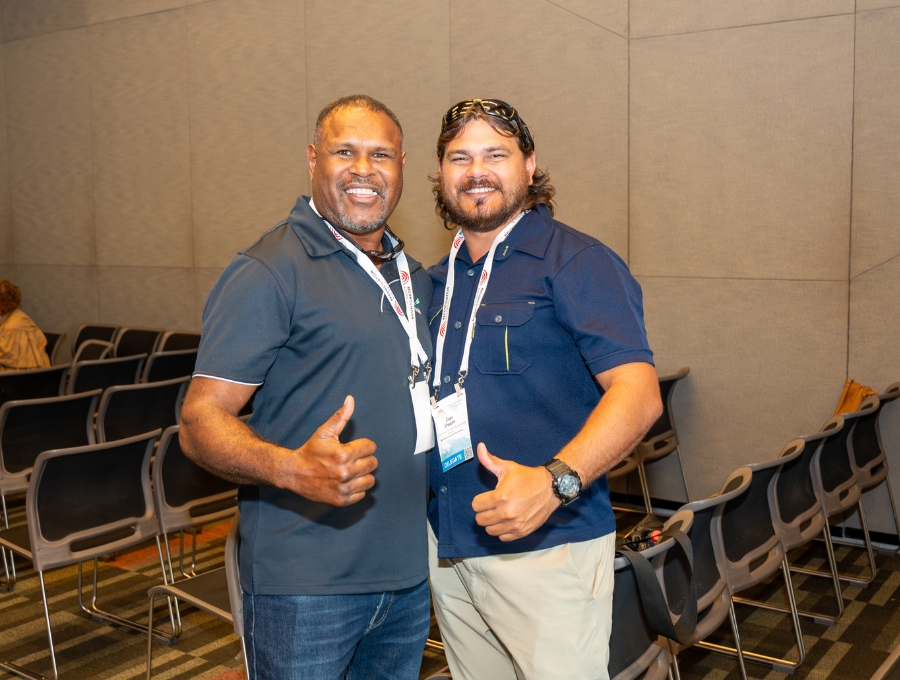 Two men smile into the camera with their thumbs up.