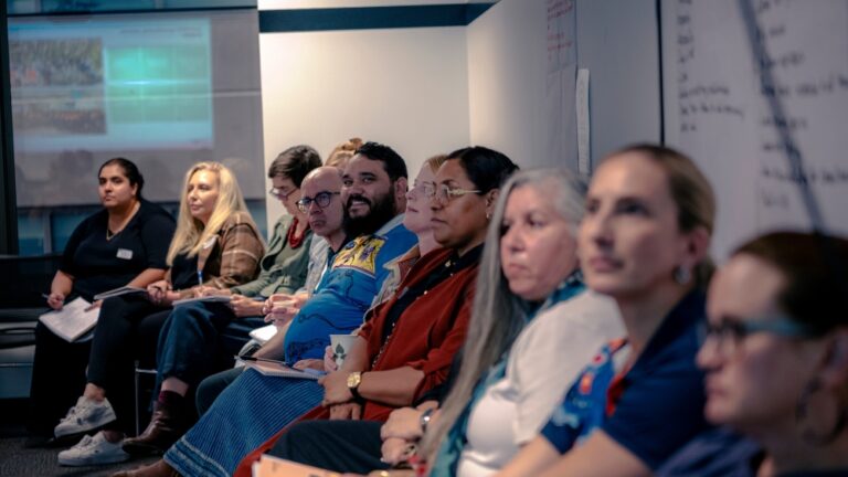 A group of people sit in a line, listening to someone out of view. A bearded man in the middle of the line smiles cheerfully into the camera.