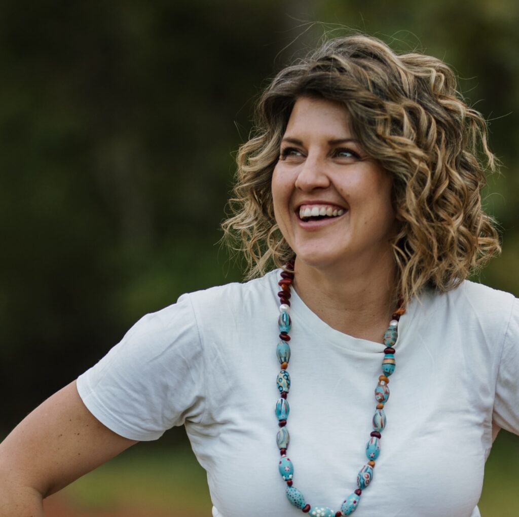 Young woman, curly hair, white t-shirt, long shell necklace, smiling off camera.