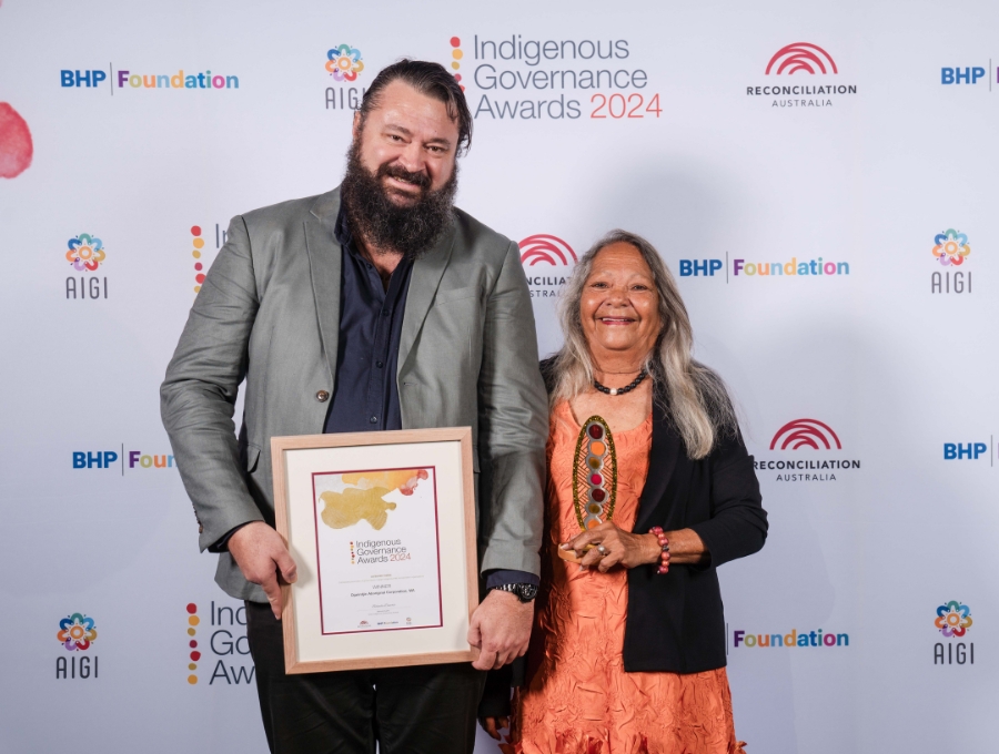 Two people stand in front of a media wall with the Reconciliation Australia red sunrise logo and the Indigenous Governance Awards 2024 logo on it. The man, taller, is holding a certificate and the woman is holding a trophy.