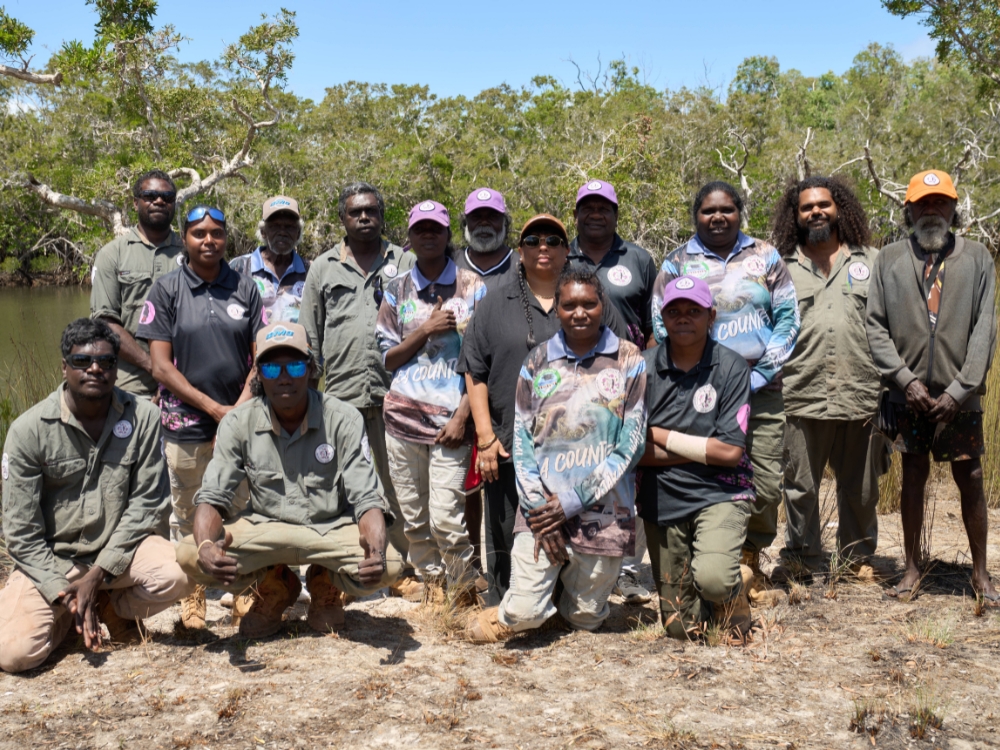 A large group of Aboriginal rangers in khaki or brightly-coloured fishing shirts, sitting, kneeling and standing on dry grass by a river.