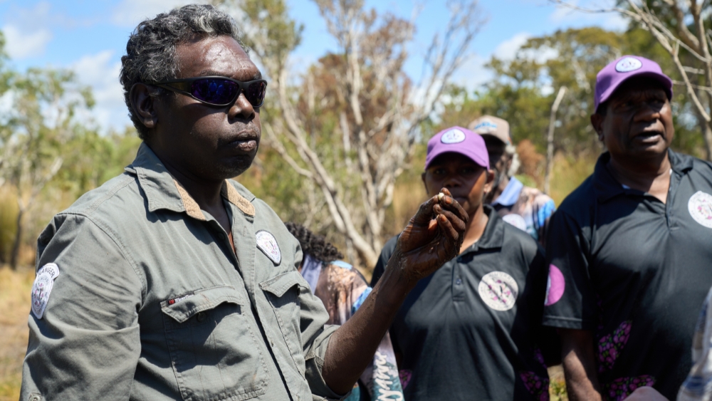 An Yolngu Aboriginal man holds a small seed, standing in a dry, scrub-like area.