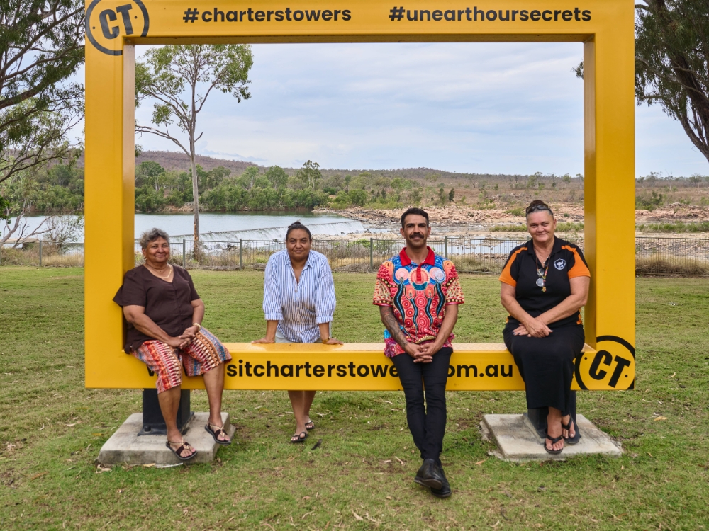 Four people sitting or leaning on a big yellow frame in a park by a body of water - the frame has the words #charterstowers and #unearthoursecrets written on it.