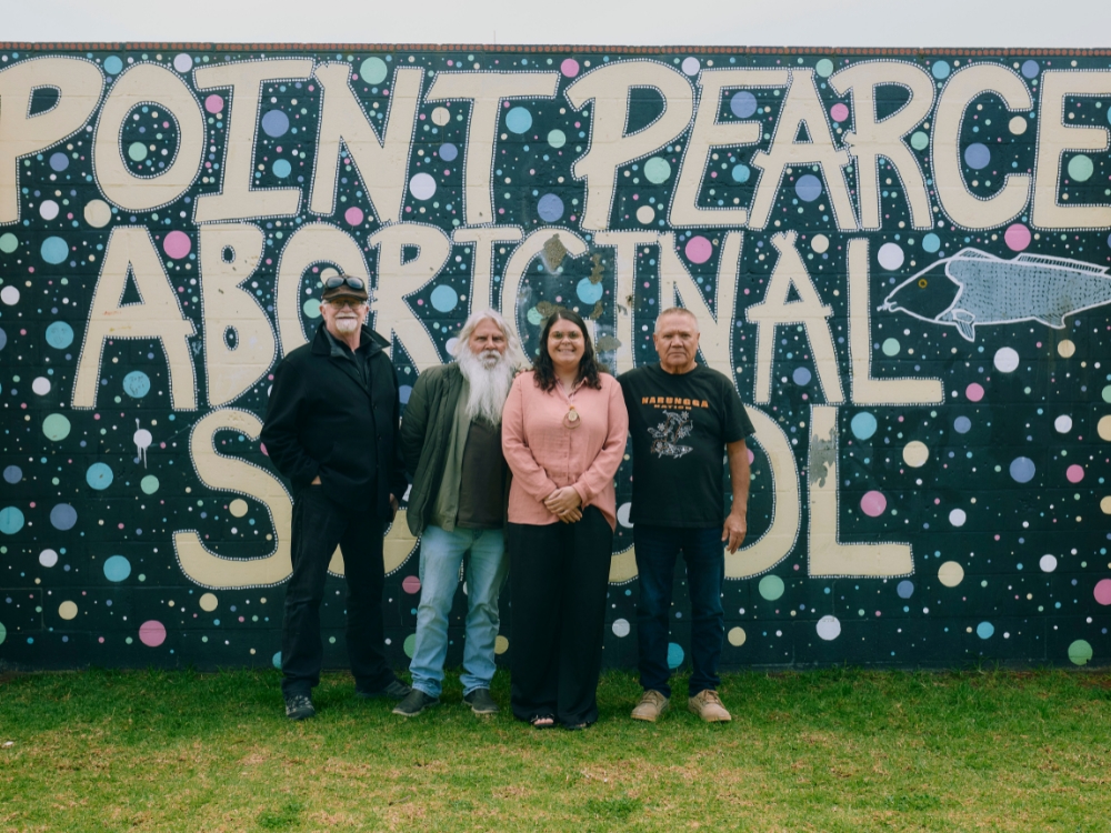 Four people standing in front a big black, white and blue mural with multicoloured dots - the white text reads 'Point Pearce Aboriginal School'