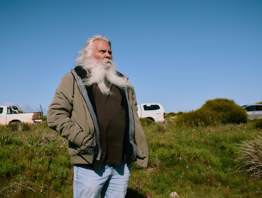 Aboriginal Elder Uncle Eddie Newchurch - long silver hair and beard - standing on open grassy plain, with the wind opening his beard into a fork.