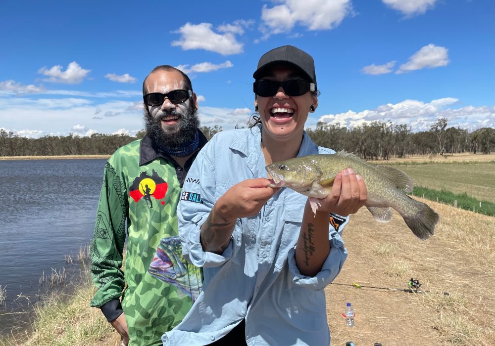 A young man and a young woman. Both are smiling and laughing. The woman is holding a fish she has just caught.