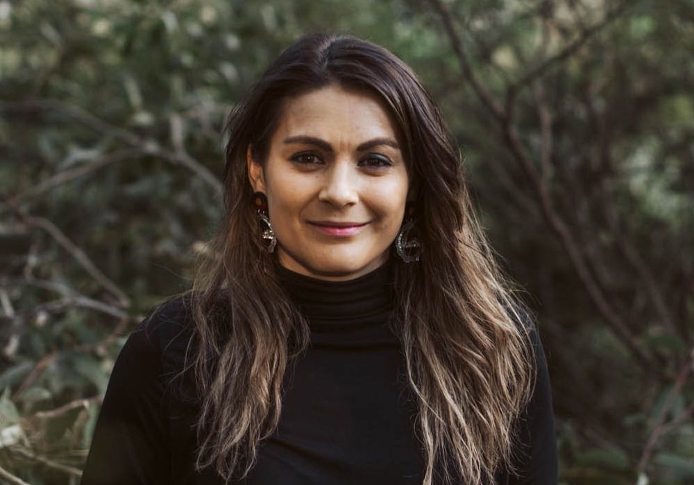 Muthi Muthi/Gunditjmara woman Jacara Egan, standing in the bush, smiling.